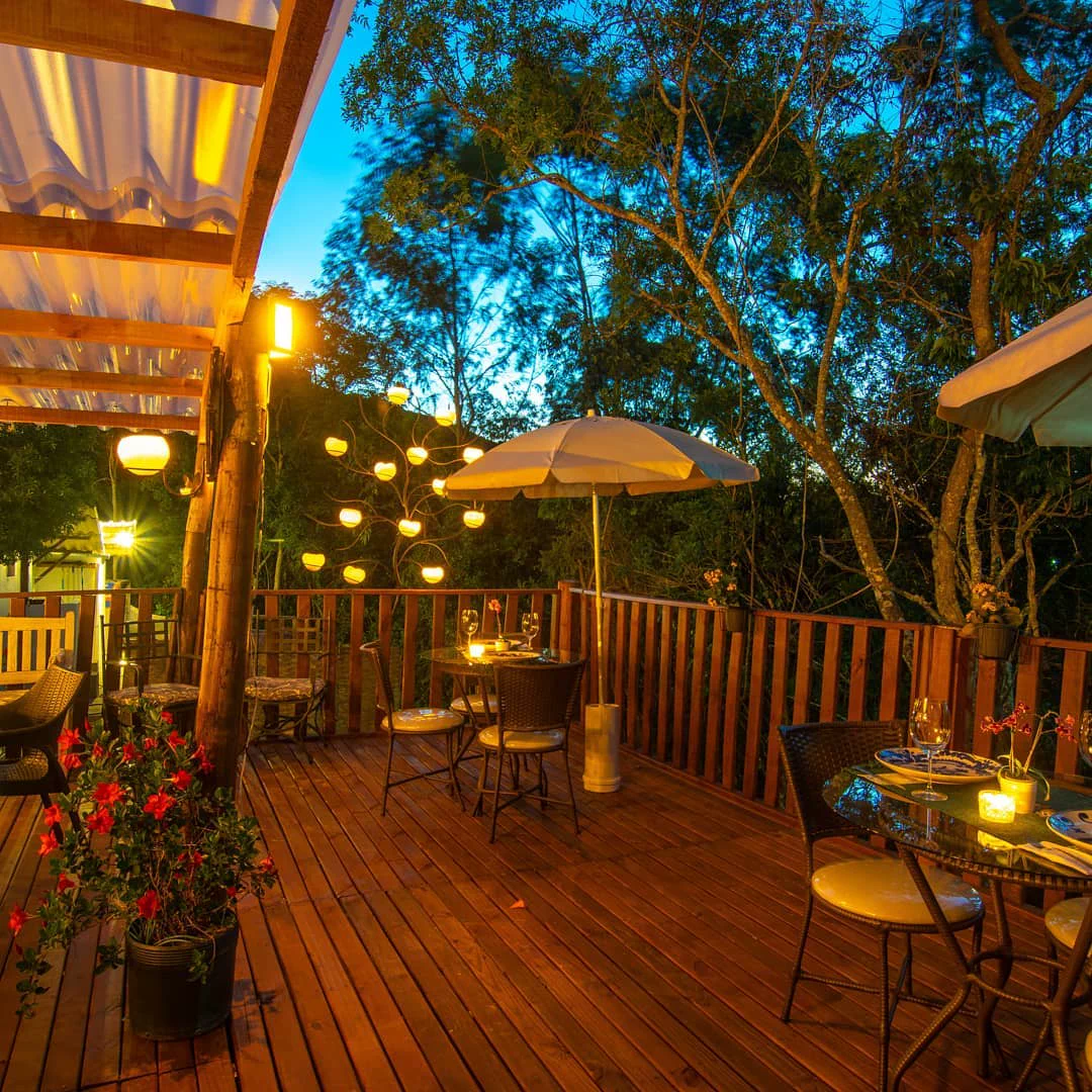 Deck de madeira ao anoitecer, cercado de árvores, com luminárias acesas e mesas postas
